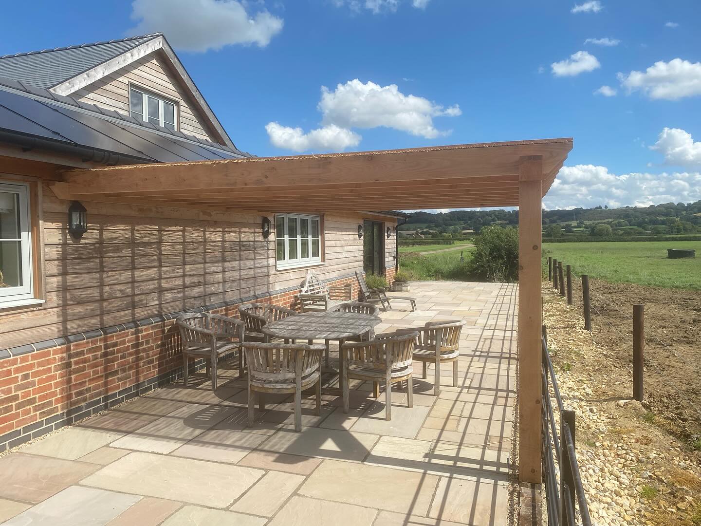 Timber and brick house with an oak pergola over a stone terrace, looking onto fields