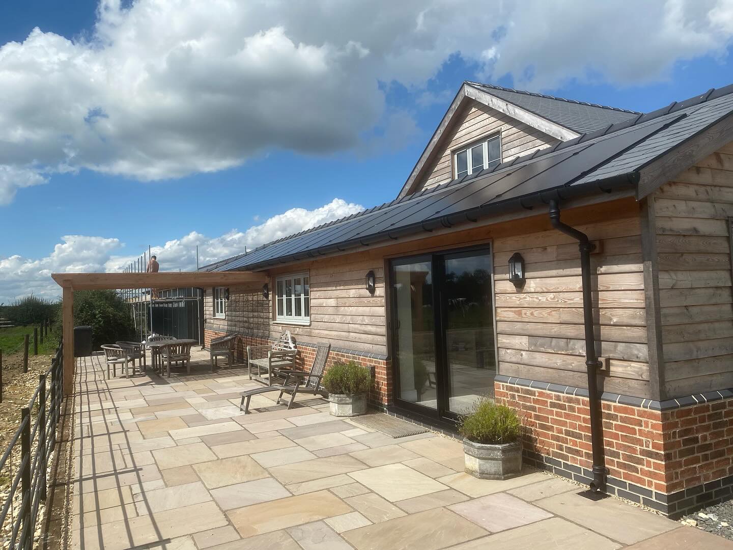 Oak-clad house with a brick base, dark tiled roof, and sliding doors onto a stone terrace